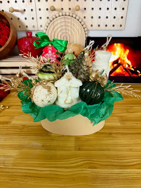 Arreglo en cesta con piezas blancas y verdes, papel verde y ramas doradas sobre una mesa de madera.