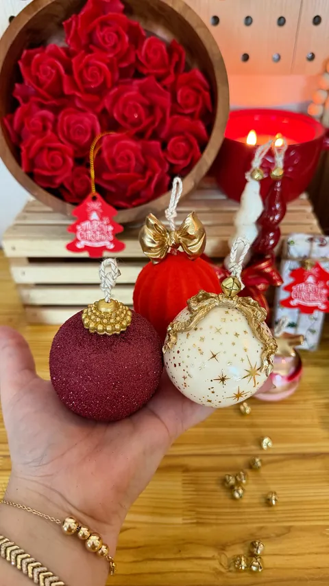 Tres esferas decorativas en tonos burdeos, rojo y blanco, con detalles y lazos dorados, sostenidas en una mano.