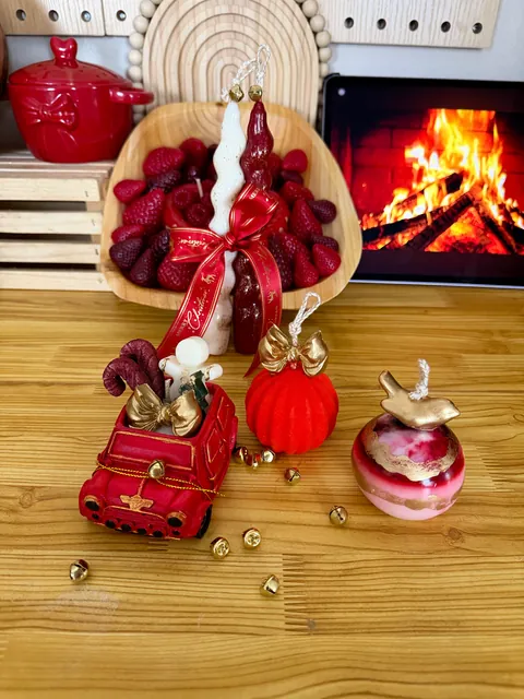 Composición sobre una mesa con velas altas blanca y roja, lazo rojo, un vehículo rojo y una esfera roja con remate dorado.